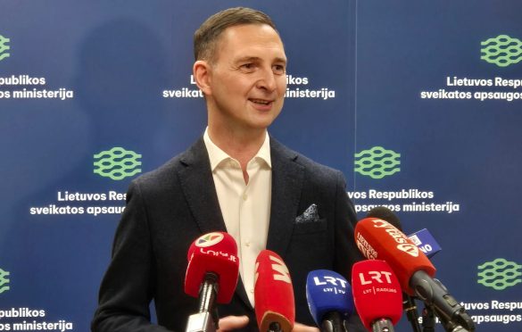 Asmuo su tamsiu švarku ir baltais marškiniais kalba spaudos konferencijoje prieš foną su užrašu „Lietuvos Respublikos sveikatos apsaugos ministerija“; matomi keli mikrofonai iš skirtingų žiniasklaidos kanalų, įskaitant LRT ir LNK.