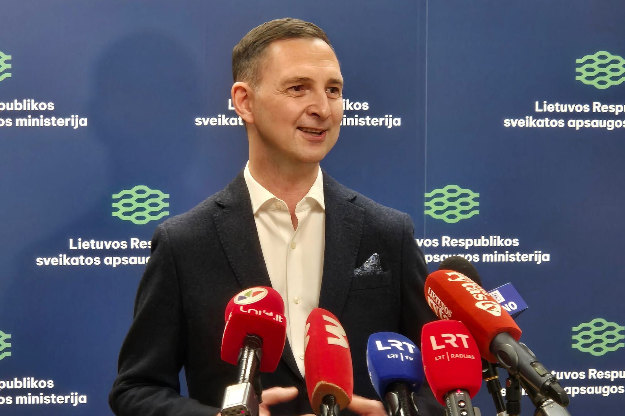 Asmuo su tamsiu švarku ir baltais marškiniais kalba spaudos konferencijoje prieš foną su užrašu „Lietuvos Respublikos sveikatos apsaugos ministerija“; matomi keli mikrofonai iš skirtingų žiniasklaidos kanalų, įskaitant LRT ir LNK.