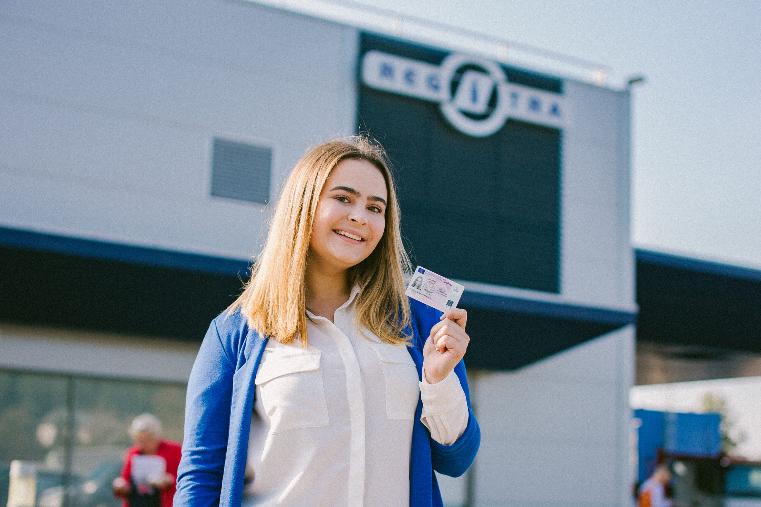 Šioje nuotraukoje matyti žmogus, stovintis priešais pastatą ir laikantis vairuotojo pažymėjimą. Ant pastato matomas užrašas „REGITRA“, kuris greičiausiai yra dalinai matomas „REGITRA“ pavadinimas.