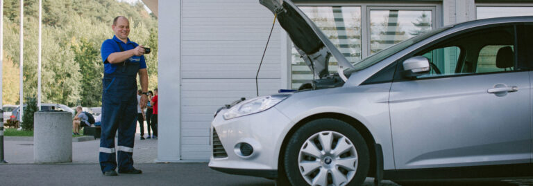 Šioje nuotraukoje matyti žmogus, vilkintis mėlyną darbo aprangą, stovintis šalia sidabrinio automobilio, kurio variklio dangtis atidarytas. Asmuo rankose laiko daiktą – galimai įrankį ar prietaisą. Fone matomas pastatas su dideliais langais, medžiai ir keli kiti žmonės.