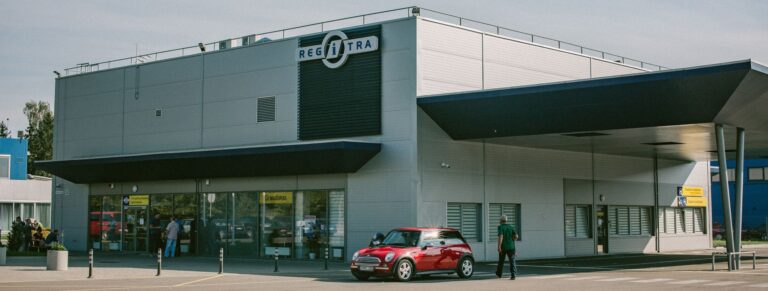 Šioje nuotraukoje matyti pastatas su užrašu „REGITRA“. Prie įėjimo stovi raudonas automobilis, netoliese eina žmogus. Pastatas turi didelius langus ir stogelį virš įėjimo, o priešais įėjimą yra stulpeliai (bollard'ai), apsaugantys nuo automobilių privažiavimo.