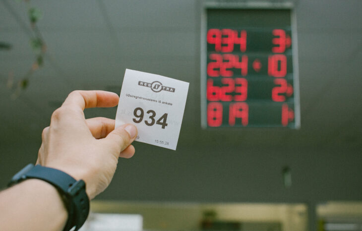 Šioje nuotraukoje matoma ranka, laikanti bilietą su numeriu „934“. Ant bilieto yra tekstas „REGITRA“ ir „Užsiregistravusiems iš anksto“, nurodantis, kad tai registracijos bilietas, skirtas lankytojams, atvykusiems į padalinį.