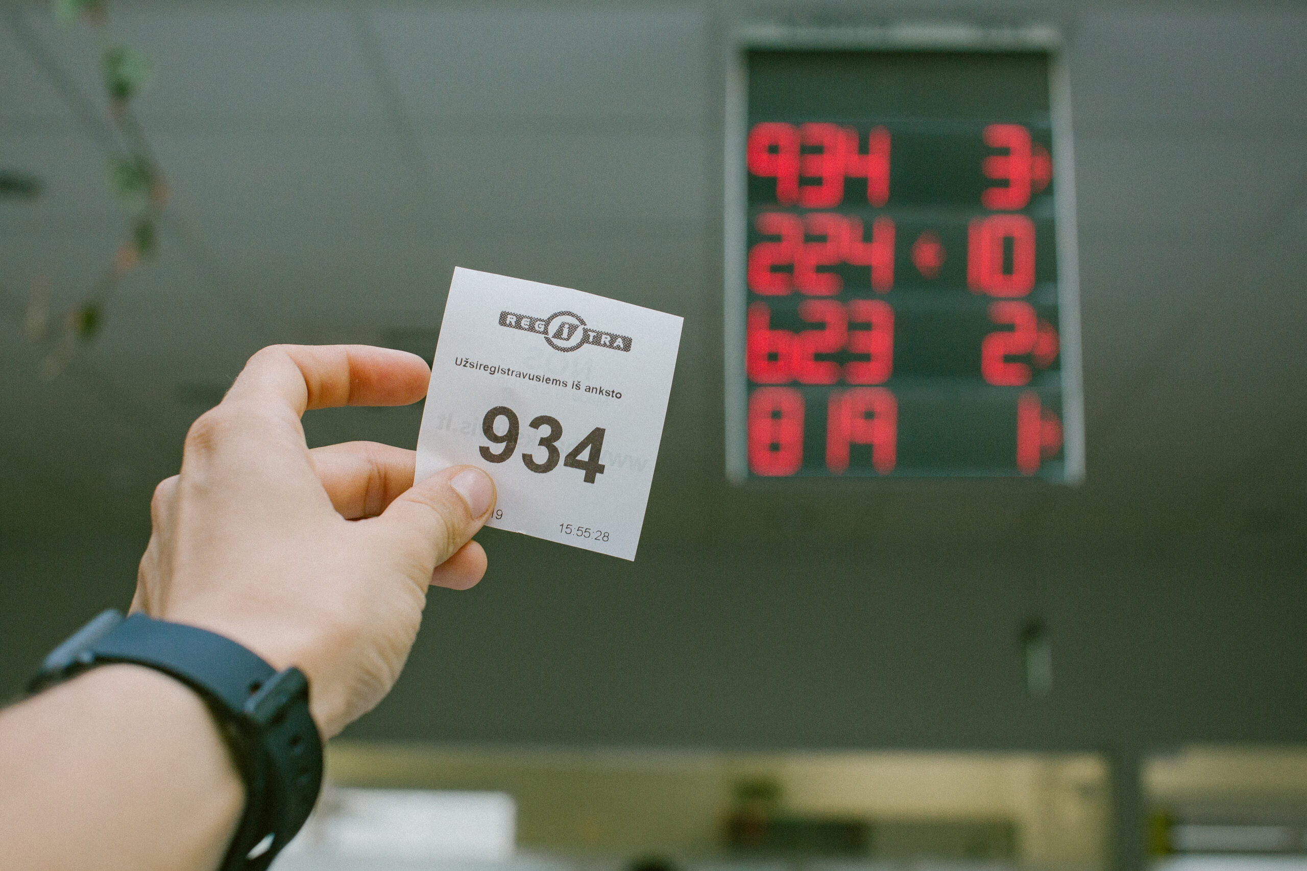 Šioje nuotraukoje matoma ranka, laikanti bilietą su numeriu „934“. Ant bilieto yra tekstas „REGITRA“ ir „Užsiregistravusiems iš anksto“, nurodantis, kad tai registracijos bilietas, skirtas lankytojams, atvykusiems į padalinį.
