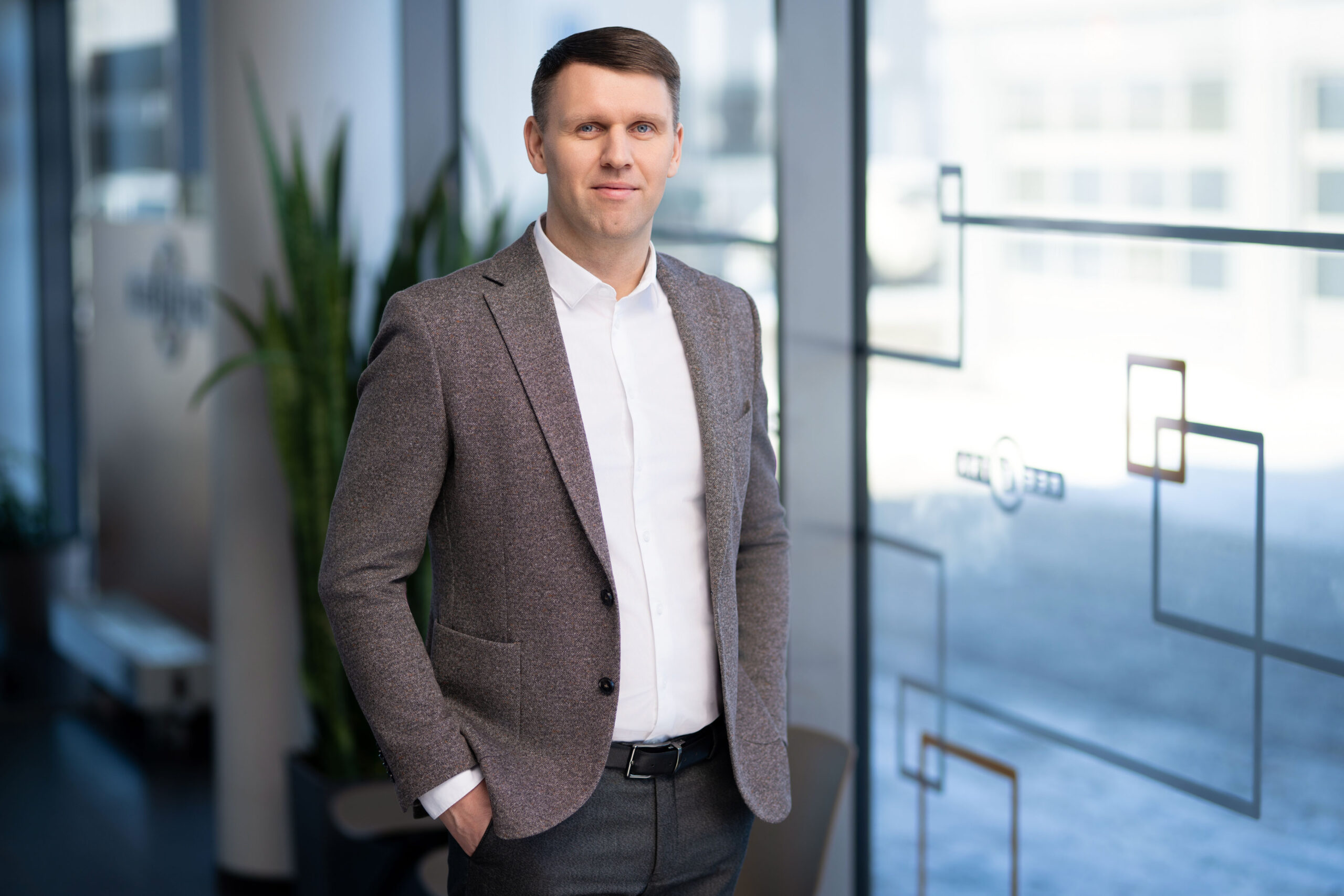 Vyras stovi su baltais marškiniais ir rudu švarku.