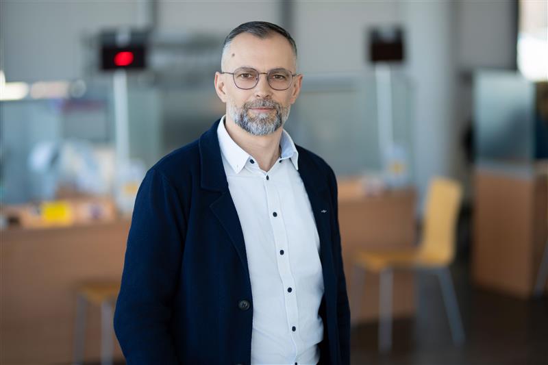 Vyras stovi su baltais marškiniais ir juodu megztiniu.