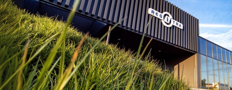 Modernus pastatas su juodu fasadu ir užrašu „REGITRA“, priekyje – žali aukšti žolės stiebai.