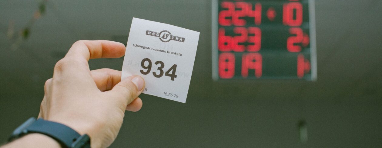 Paveikslėlyje matoma ranka, laikanti bilietėlį su numeriu „934“. Ant bilieto taip pat yra užrašai „REGITRA“ ir „Užsiregistravusiems iš anksto“. Fone matomas skaitmeninis ekranas, kuriame rodomi šie numeriai: 224 – 0 823 – 2 819 – 1