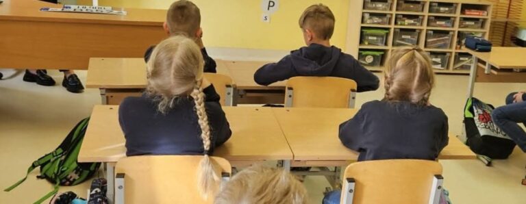 Mokyklos klasė su penkiais vaikais, sėdinčiais prie stalų nugara į kamerą; vaikai vilki tamsius drabužius, šalia jų – kuprinės, fone matyti mokytojo stalas ir lentynos su mokymo priemonėmis.