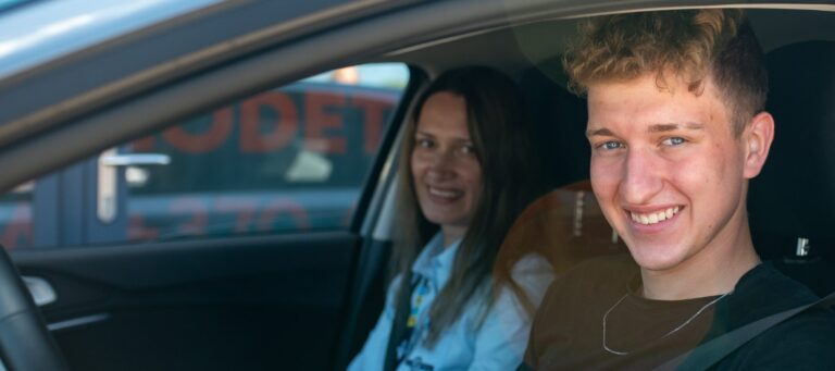 Jaunas vairuotojas sėdi prie mašinos vairo šalia sėdi moteris, abu šypsosi