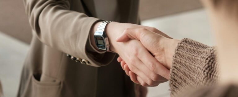 Stambiu planu užfiksuotas dviejų žmonių rankų paspaudimas – vienas vilki smėlio spalvos švarką ir dėvi skaitmeninį laikrodį, kitas vilki megztinį.