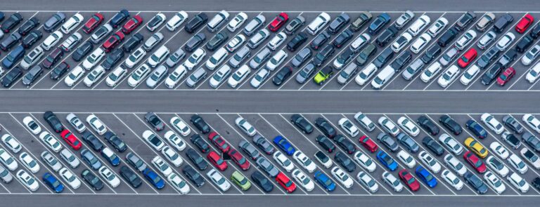 Aukštuminis vaizdas į didelę automobilių stovėjimo aikštelę su tvarkingai išrikiuotomis transporto priemonėmis dviejose sekcijose, atskirtose centrine juosta. Matomi įvairių spalvų sedanai, visureigiai ir sunkvežimiai