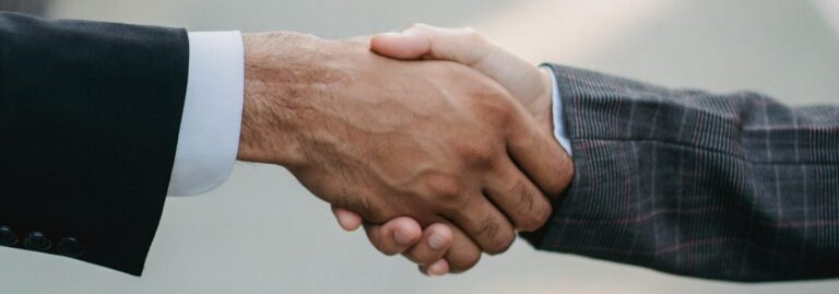 Stambiu planu užfiksuotas dviejų žmonių rankų paspaudimas – vienas vilki juodą kostiumą su baltais marškiniais, kitas – pilką languotą kostiumą.