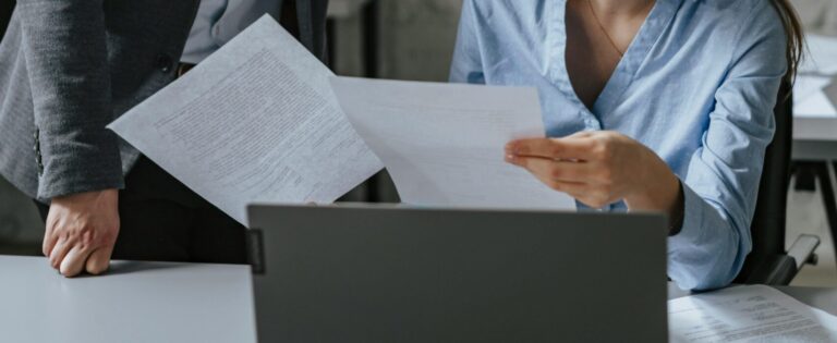 Du žmonės profesionalioje aplinkoje – vienas stovi laikydamas dokumentą, kitas sėdi prie stalo su nešiojamuoju kompiuteriu ir peržiūri dokumentus.