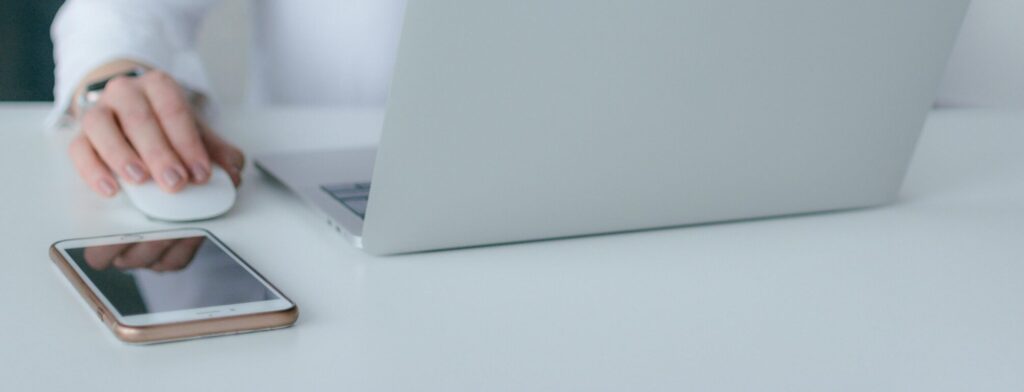 žmogus ieško informacijos internete, ranka padėta ant kompiuterio pelės, nešiojamas kompiuteris ant stalo 