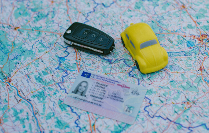 Žemėlapio fone padėtas automobilio raktas, geltonas žaislinis automobilis ir Lietuvos vairuotojo pažymėjimas
