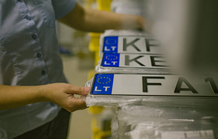 Asmuo tvarko krūvą Europos Sąjungos stiliaus automobilių numerių su mėlynu ES simboliu ir žyma „LT“. Matomi numeriai: „KP“, „KE“, „FA1“.