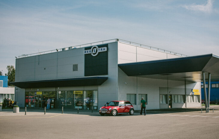 Modernus pastatas su užrašu „REGITRA“, prie įėjimo stovi raudonas automobilis ir keli žmonės. Pastatas turi didelius langus ir stogelį virš įėjimo.