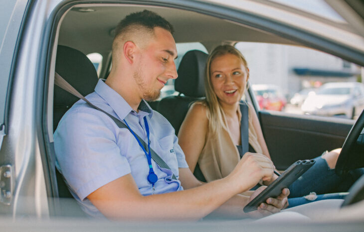 Du žmonės sėdi automobilyje – kairėje esantis asmuo su šviesiai mėlynais marškiniais ir kortele ant kaklo laiko planšetę, o dešinėje esantis asmuo su berankove palaidine žiūri į įrenginį. Pro langus matyti stovintys automobiliai