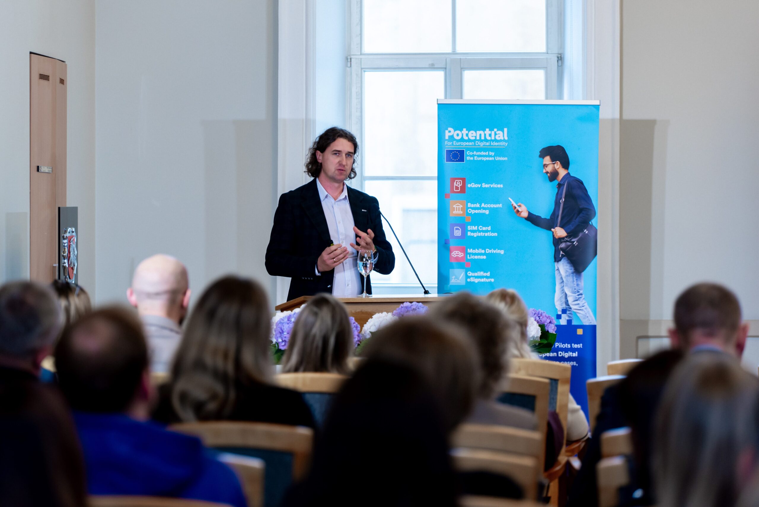 Vyras su švarku kalba prie tribūnos konferencijoje