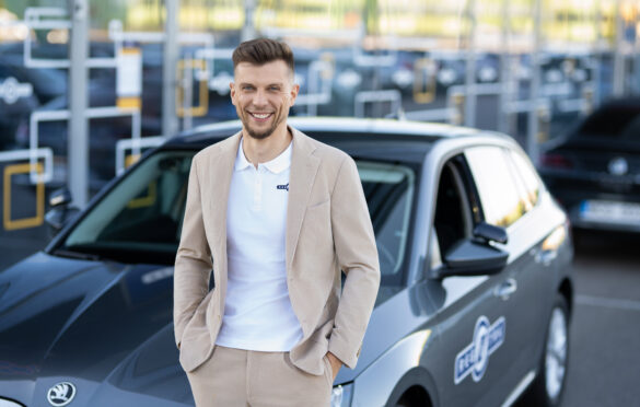 Vyras stovi su šviesiu kostiumu atsirėmęs į automobilį