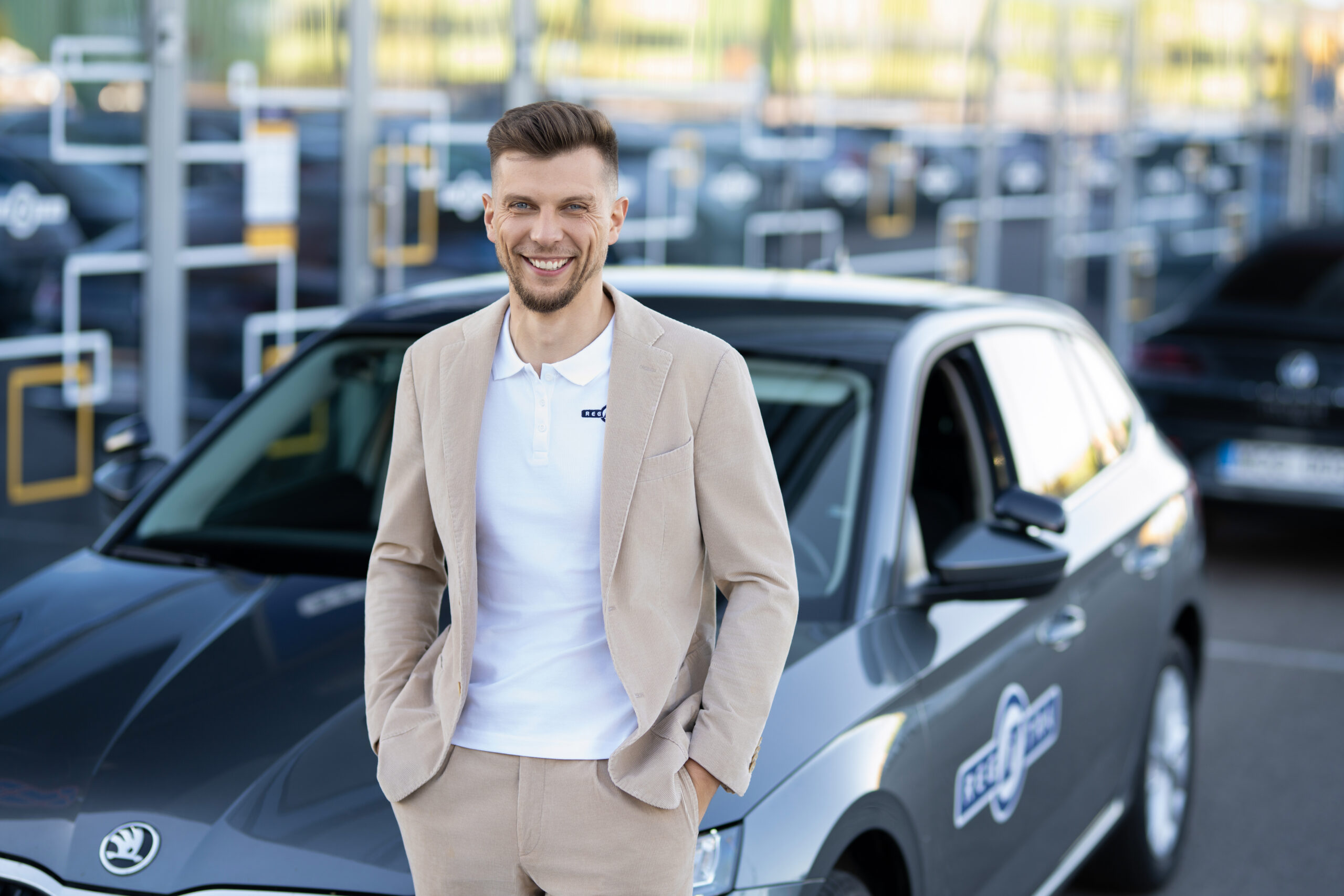 Vyras stovi su šviesiu kostiumu atsirėmęs į automobilį