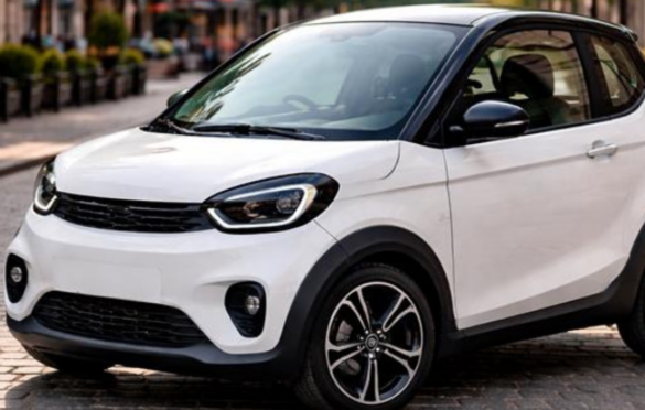 Balta kompaktiška miesto automobilio tipo transporto priemonė stovi ant grindinio miesto gatvėje; fonas šiek tiek neryškus, matyti pastatai ir želdiniai.