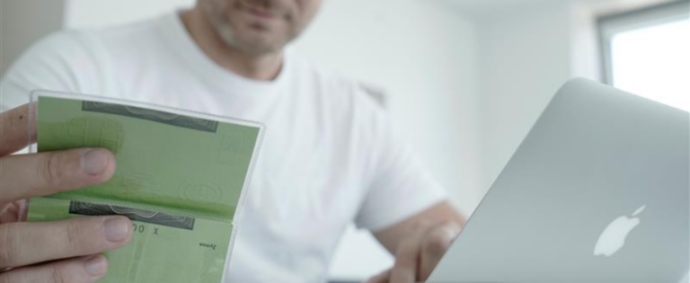 Vyras sėdi prie nešiojamojo kompiuterio ir rankoje laiko žalią transporto priemonės registracijos liudijimą.